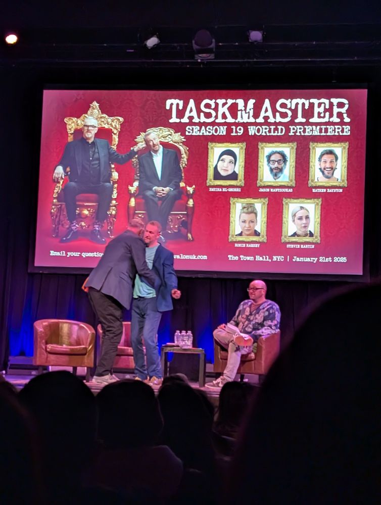 Alex looking unthrilled but accepting of his fate as Greg begins to grab around his waist to pick him up, as requested by an audience member 