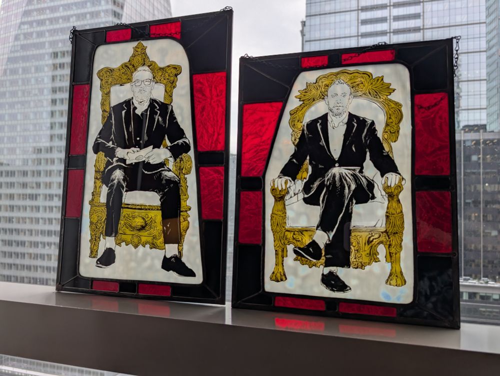 Stained glass portraits of Greg Davies and Alex Horne that are leaning against a window with a view of NYC buildings as backdrop 