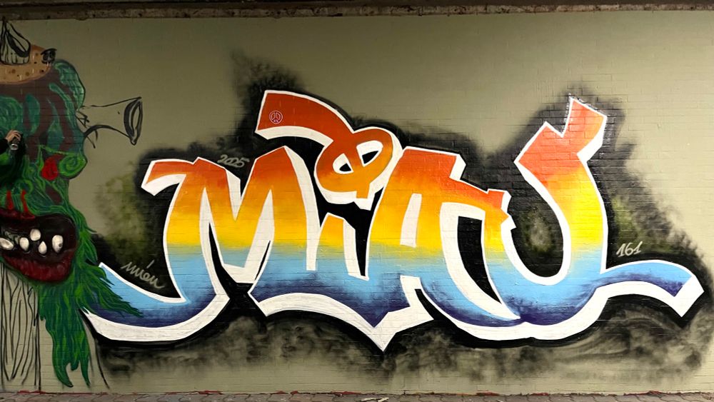 Bunte Wandgestaltung, Graffiti aus der Buchstabenkombination MIAU, die Farbgestaltung der Buchstaben entspricht Farbverläufen, horizontal von unten nach oben geschichtet, von lila zu hellblau zu gelb zu orange zu rotorange, die Outlines und der 3d Effekt sind in weiß, der Hintergrund schwarz genebelt zur sonst grün gestrichenen Wand.
