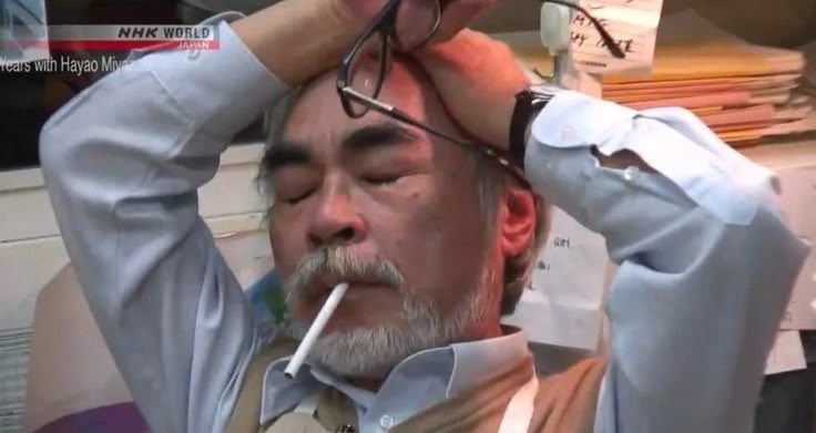 Hayao Miyazaki looking exhausted leaning back in his chair with his eyes closed, cigarette in mouth, and both hands on his head one of which is holding his glasses. 