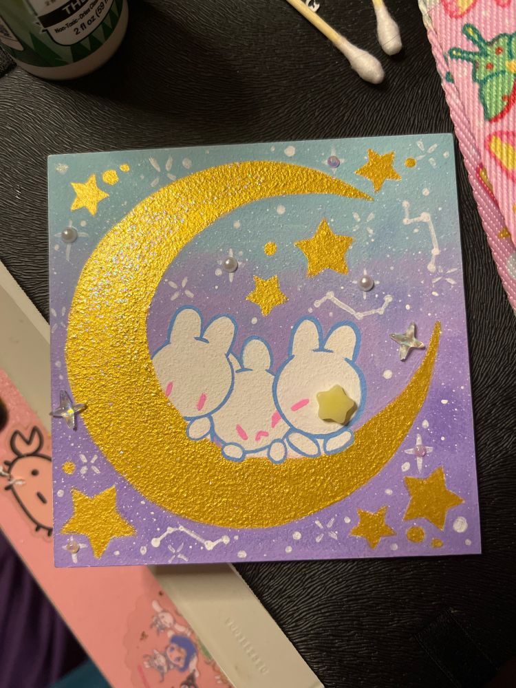 A small painting of three white bunnies crowded together on a moon. The painting is mostly purples/blues with the moon and stars being gold. There are some 3D details with decorative cabochons. 