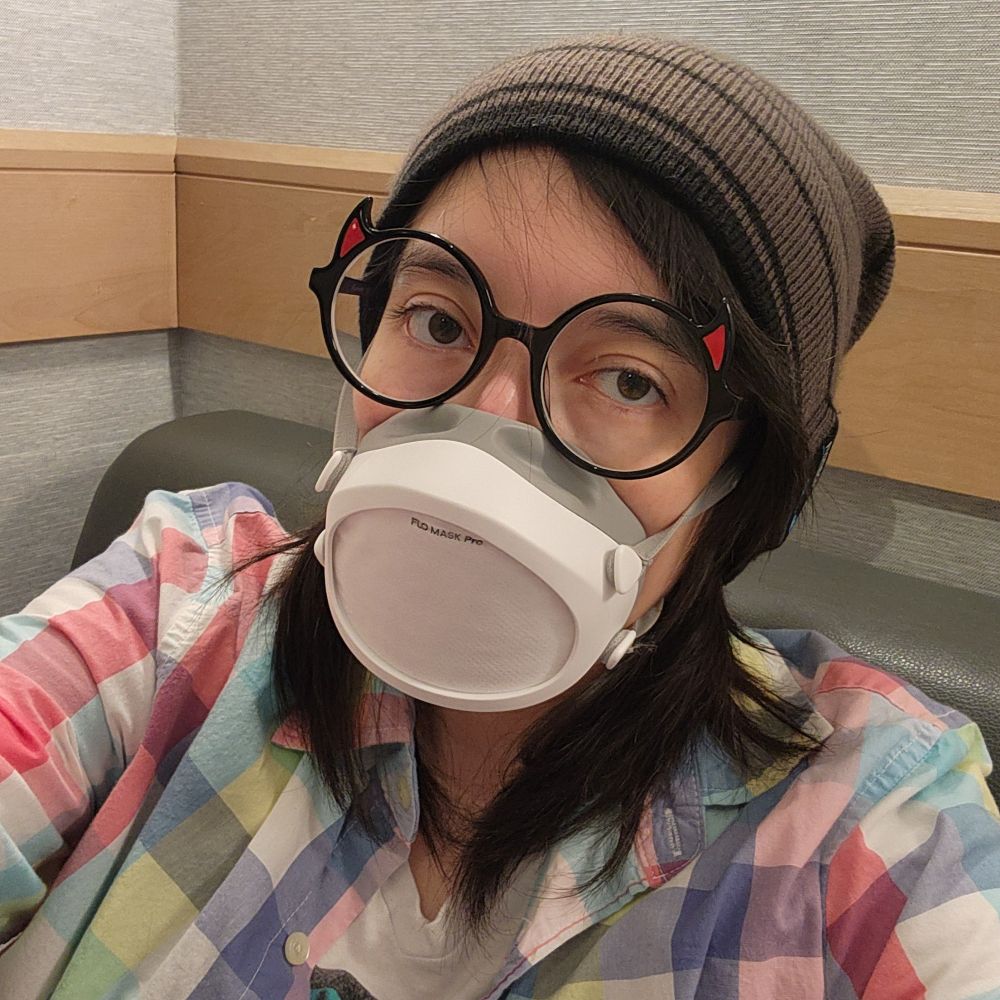 Khaos/Baires is wearing black round glasses with curved red triangle tips that are at the top corner of each circle frame that makes it look like winged eyeliner, a gray with black striped beanie, a flo mask with a pro filter, and a plaid button up.
