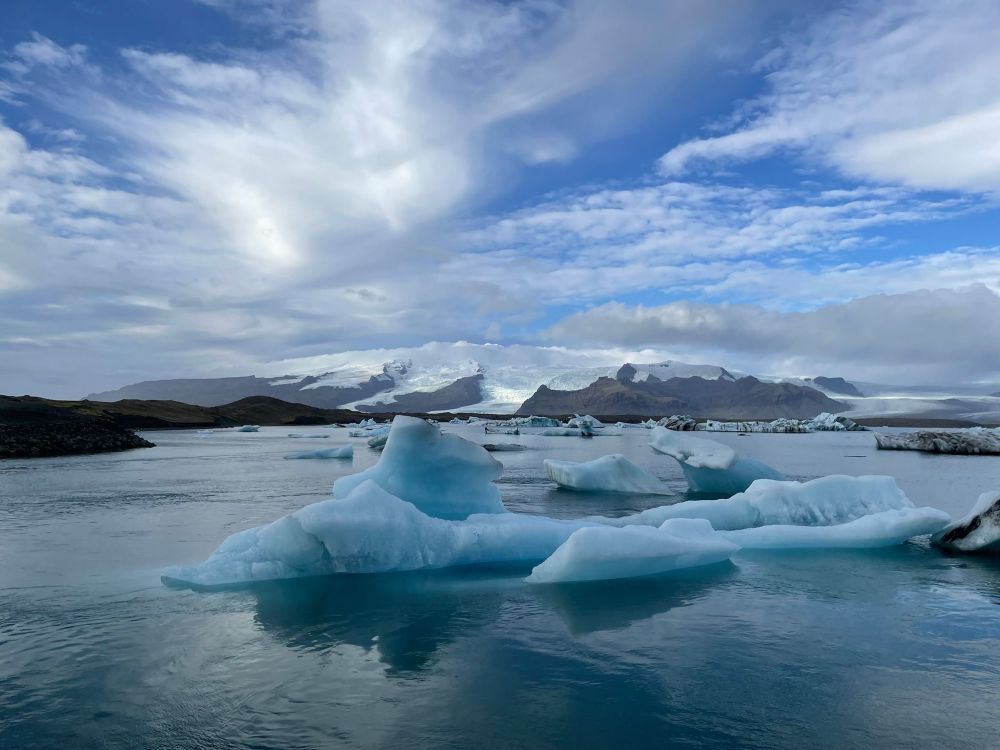 Island. Blauer Himmel, mit weißen Wolken über einer Gletscherlagune. Darin treiben Eisschollen, die auf dem Foto hellblau scheinen.
