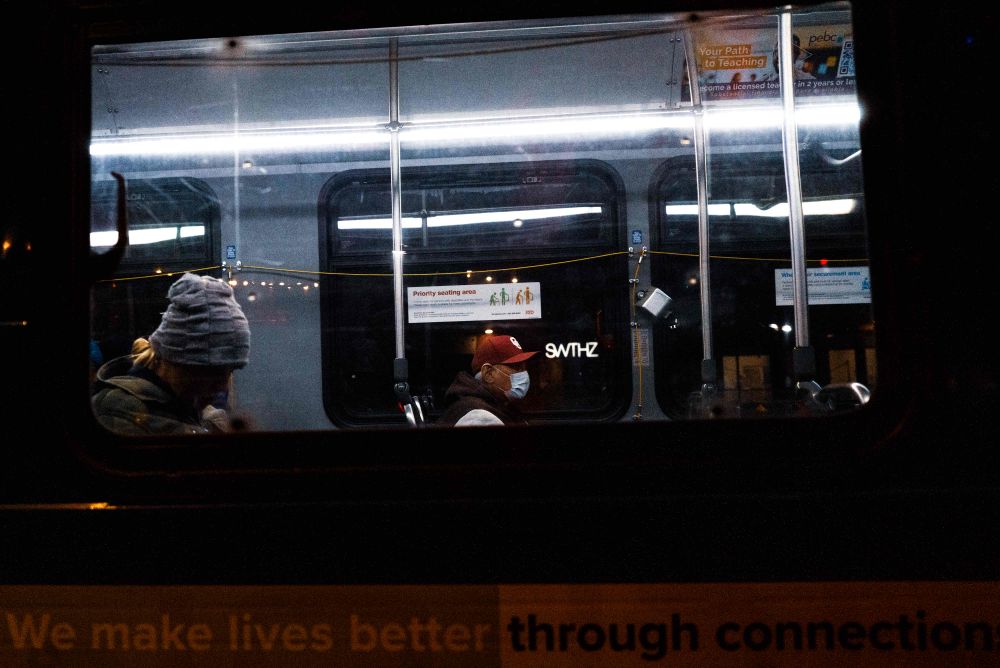 A transit bus at night, lit up inside with passengers keeeping to themselves. Along the side of the bus reads “we make lives better through connection”