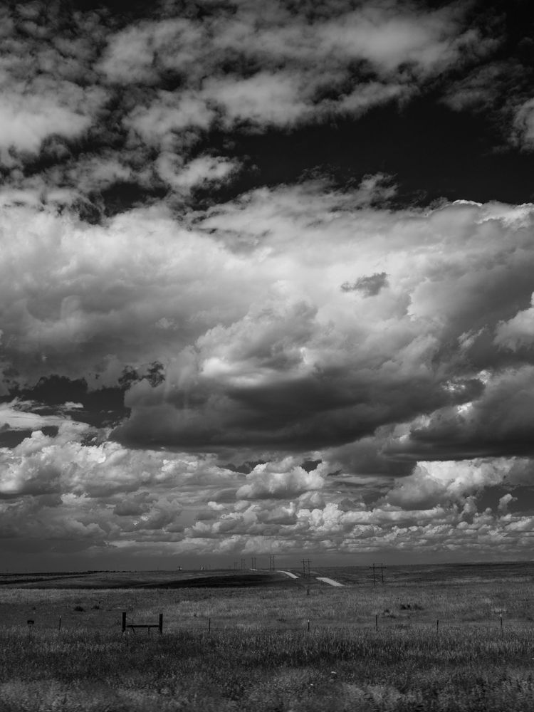 The sky is filled with large, puffy clouds with dark undersides. Looking over the plains, you can see a small road rolling over the hills. 