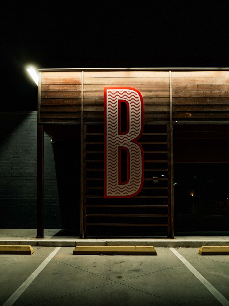 A wood-sided building stands in front of a brick wall painted black. There is a letter B on the building with a wavy pattern in the letter, and there are cuts in the concrete in the parking lot in the foreground that create a crisscross shape