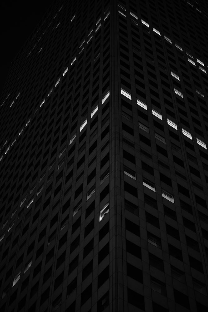 Looking up the corner of a high rise at night, most windows are dark but a few have lights on