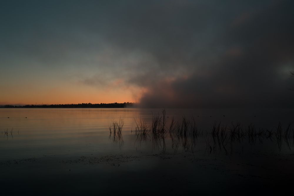 Bright orange light appears over the horizon with fog breaking up over a lake