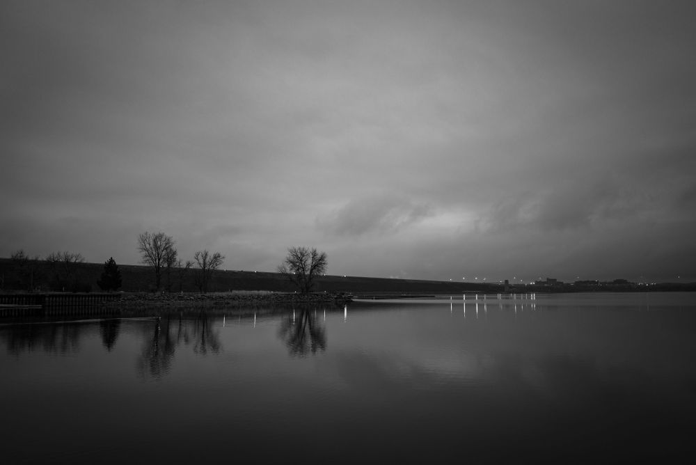 Gray skies over Dam Road of Cherry Creek Reservoir in Denver. Commuter headlights are bright, reflecting off the water