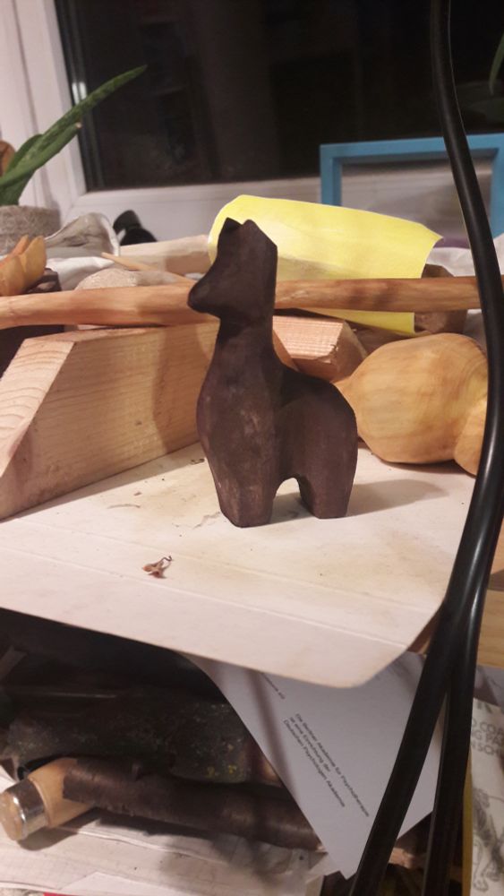 A simplified dark carved lama in a chaotic evironment with tools and other carvings in the background