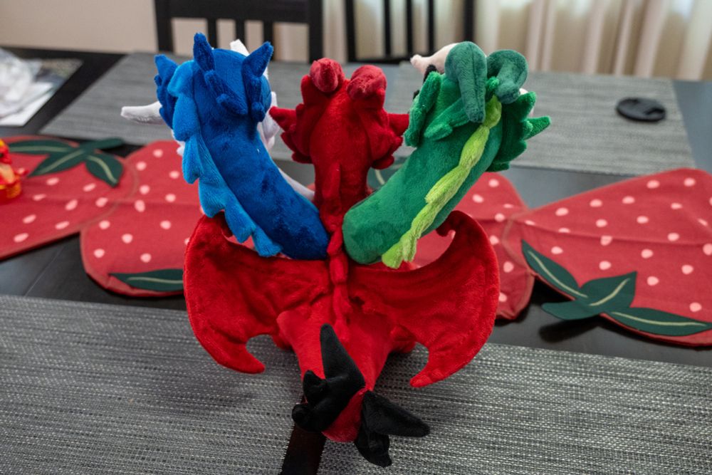 a back view of a five headed dragon plush. the wings and black-spiked tail are visible, but white and black heads are blocked from view by the blue and green heads.