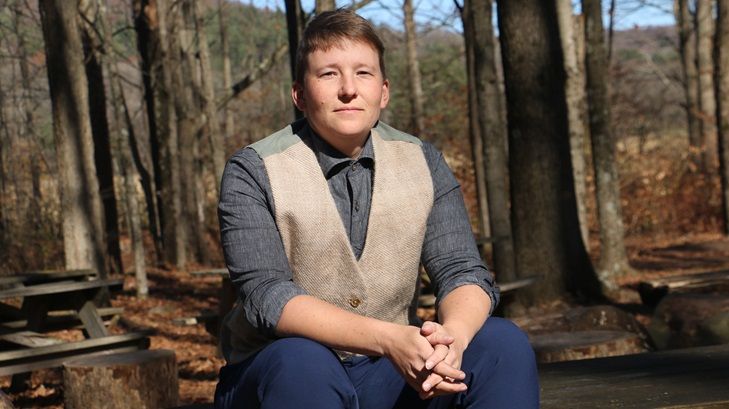 A person is sitting on a picnic table with their forearms resting on their knees, weaving a grey buttonup shirt and the tan waistcoat.