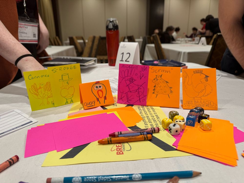 A line of simple drawings of eldritch chicken characters on index cards atop a table with scattered game supplies