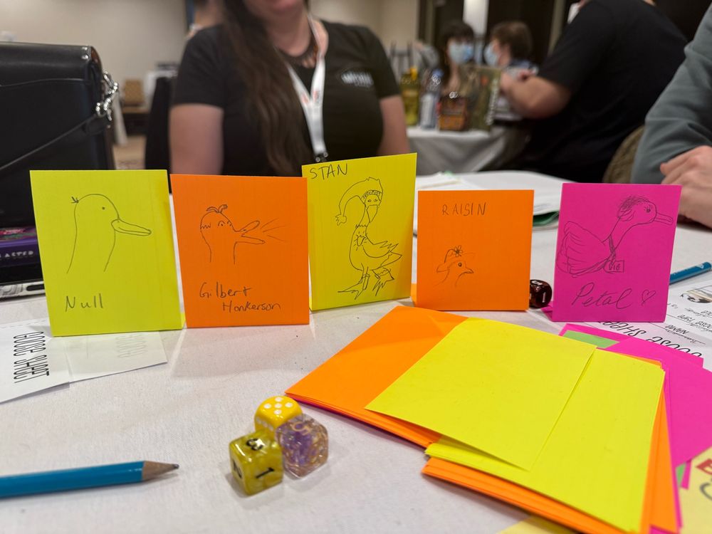 A line of simple drawings of silly geese characters on index cards atop a table with scattered game supplies