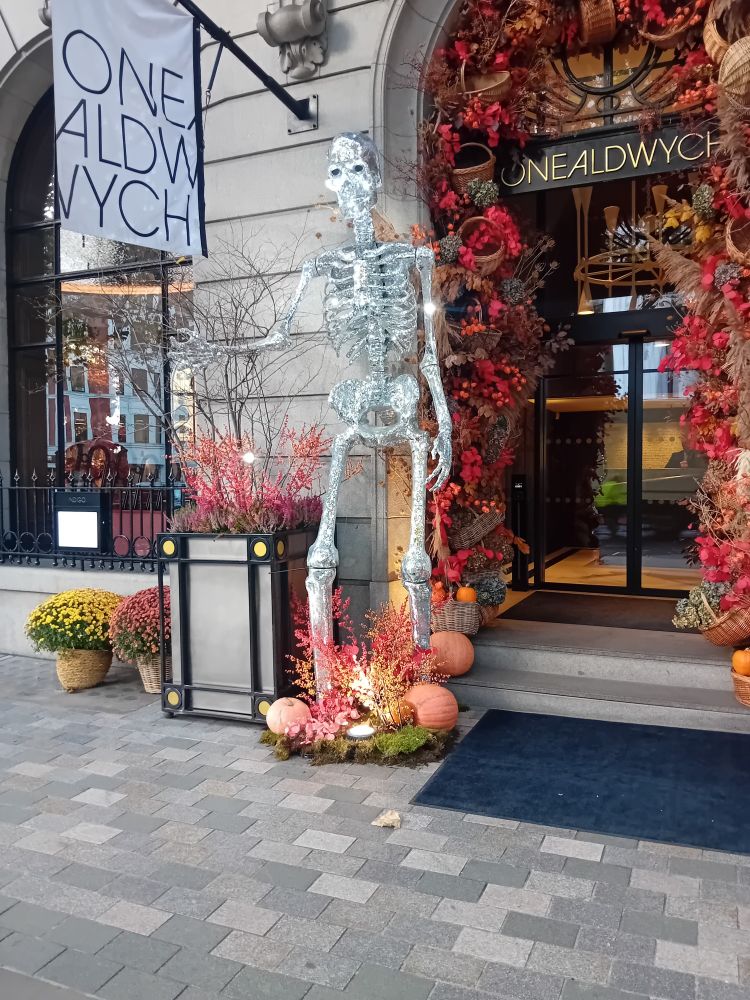 A larger than life silver skeleton at a doorway. The skeleton has pumpkins at their feet. The doorway is surrounded with an arch of pumpkins and autumn leaves.