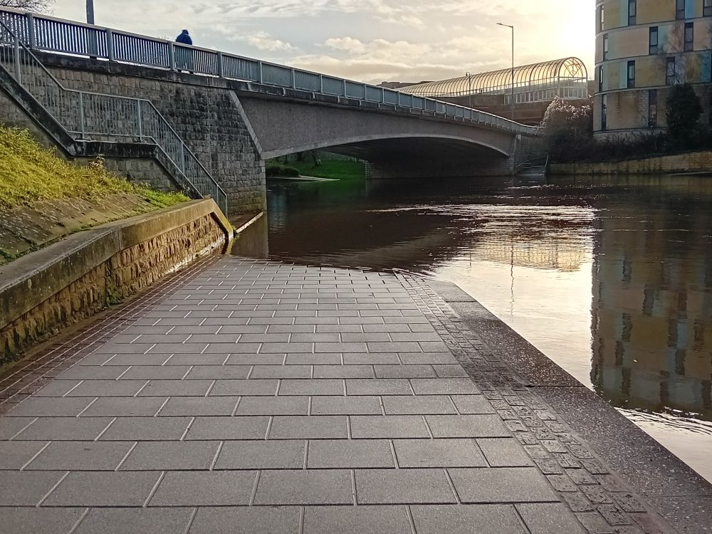 View along a path which slopes down under a bridge. The path is blocked by flooding.