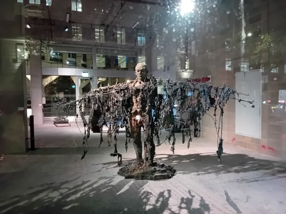 A statue of a human figure with arms stretched out to the sides. The arms and body are draped with tattered rags.