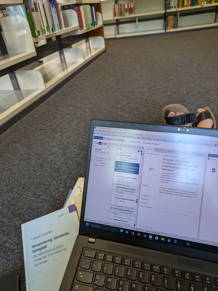 Ein Laptop, Bücher und Füße auf dem grauen Fußboden einer Bibliothek. Im Hintergrund Bücherregale.