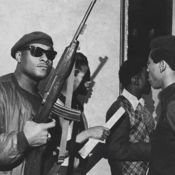 Fotografia em preto e branco de membros do Partido dos Panteras Negras em uma manifestação dentro do Capitólio da Califórnia, em Sacramento, no ano de 1967. No primeiro plano, um homem negro, vestindo boina, jaqueta de couro e óculos escuros, segura um rifle enquanto olha diretamente para a câmera. Outros membros do grupo, também armados, aparecem ao fundo, interagindo ou observando. Essa manifestação foi organizada para protestar contra o Mulford Act, uma proposta de lei que visava restringir o porte aberto de armas, algo visto pelos Panteras Negras como um ataque ao seu direito de autodefesa e à luta contra a brutalidade policial.