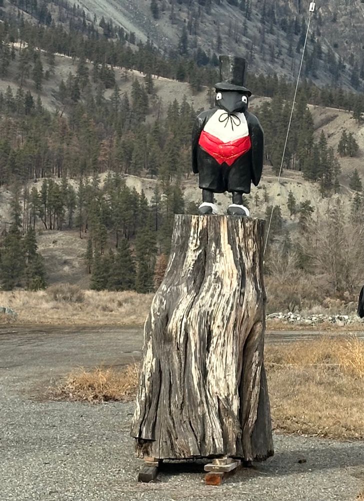 A statue of a penguin wearing a top hat standing on a chunk of wood. 