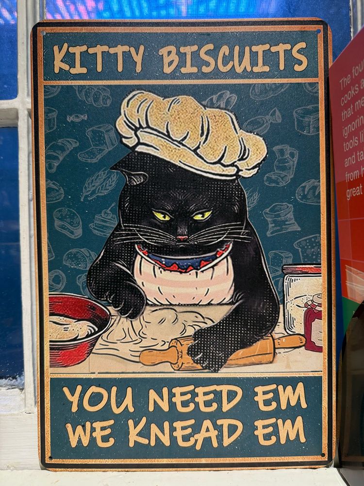 A metal poster of a black cat wearing a baker’s hat and kneading biscuit dough, text reads “Kitty Biscuits: You Need Em We Knead Em”
