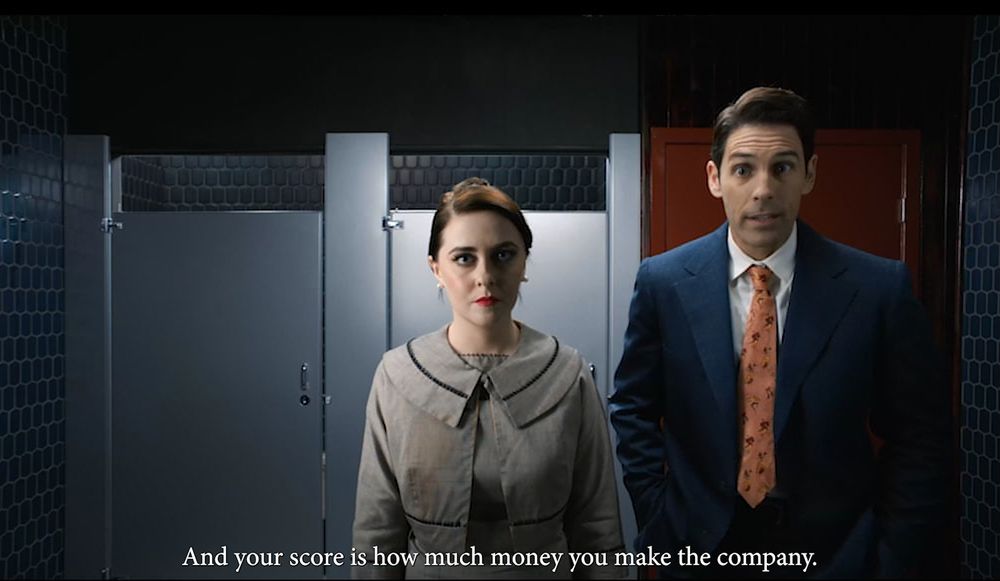 A scene in what appears to be a corporate bathroom or locker room. A woman in a vintage-style grey dress with a large collar stands next to a man in a blue suit with an orange tie. The woman has pale skin, dark hair, and red lipstick, while the man has a somewhat skeptical expression. White subtitles at the bottom read 'And your score is how much money you make the company.