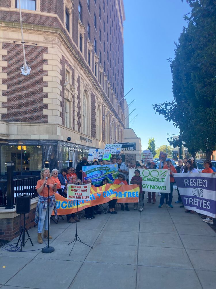 An advocate speaks while others hold banners and signs against nuclear, for renewable energy, and respecting treaties with Indigenous Nations