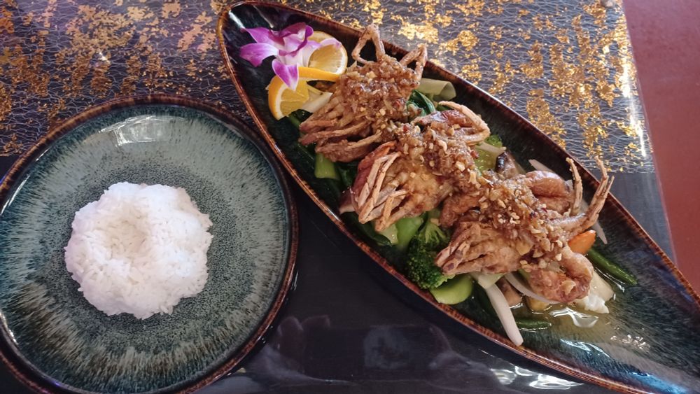 Two plates (on a black tablecloth with gold-colored accents). The small, round, plate on the left has white rice in the center, that has been in a flower-shaped mold. The larger, elliptical plate on the right has an orchid blossom and orange slice on one end, the rest of the plate is taken up by 3 softshell crabs arranged on a bed of vegetables (the vegetables are mostly green). The crabs are topped with minced garlic. 