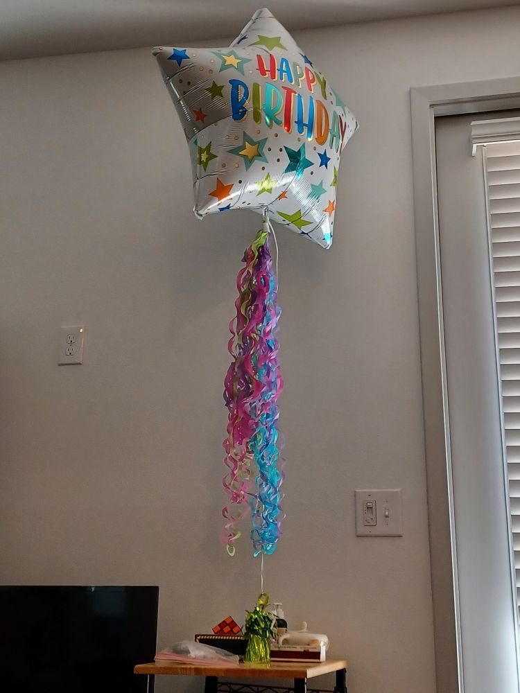 Large mylar balloon, star-shaped and with star print, that includes the words "HAPPY BIRTHDAY." Has curly pink, green, purple, and blue paper streamers attached, and is tied to a shiny green balloon weight. The ballon weight is on a small table with a few other objects including a Rubix cube and a sugar bowl. The background shows part of a flat-screen TV, a blank white wall, and a door frame with window blinds. 