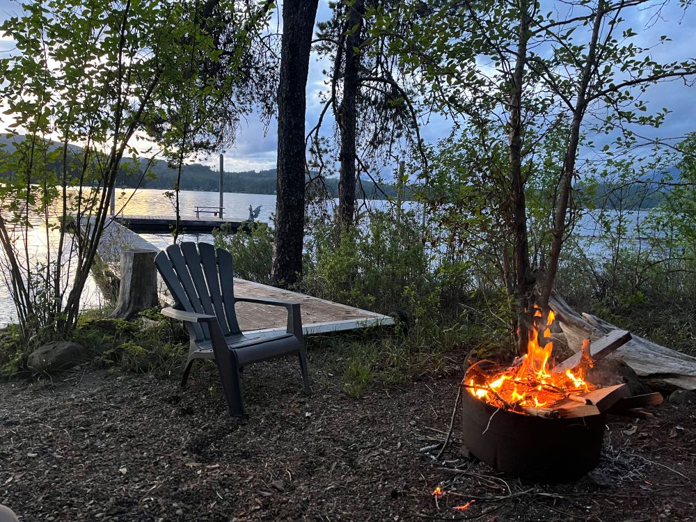 View through trees to a lake with overcast evening skies and calm water. Dock in middle ground, and in foreground a green plastic camp chair and a fire ring with small fire burning.