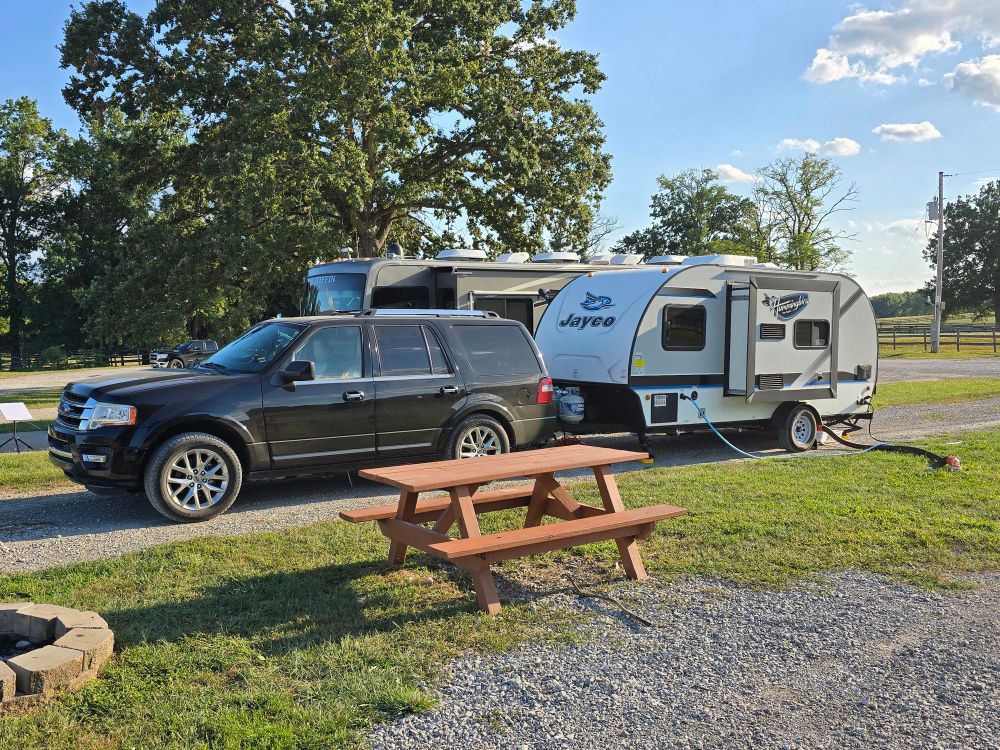 Ford Expedition hooked up to a Jayco Hummingbird 17rb travel trailer at an RV park with a picnic table and fireplace in the foreground.