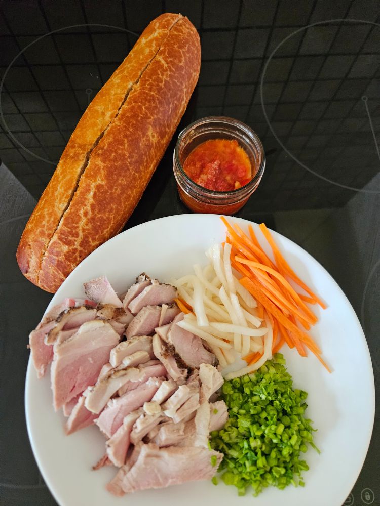 Ingredients clockwise from top left of a tiger roll, a pot of homemade chilli jam, a round white plate with slices of roast pork, matchsticks of pickled carrot & daikon, chopped coriander stems.