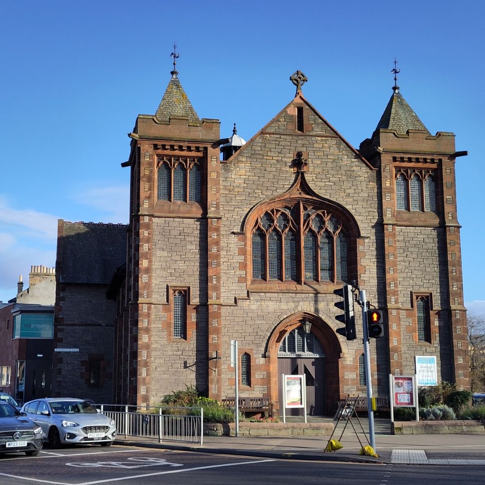 A photo of the Meadowbank and Willowbrae church building on a sunny day. It is right next to a pedestrian crossing. The building is stone with two turrets. The modern extension has a large "Try Praying" graphic completely covering the windows. 