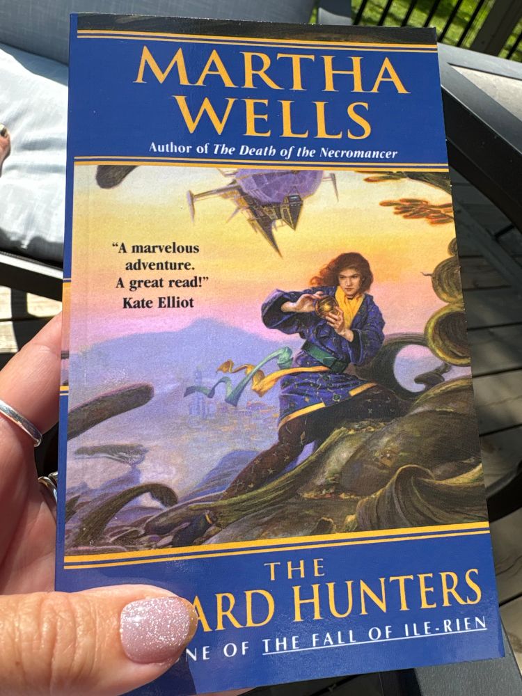 A hand holding up the paperback version of the amazing Martha Wells’ book “The Wizard Hunters”