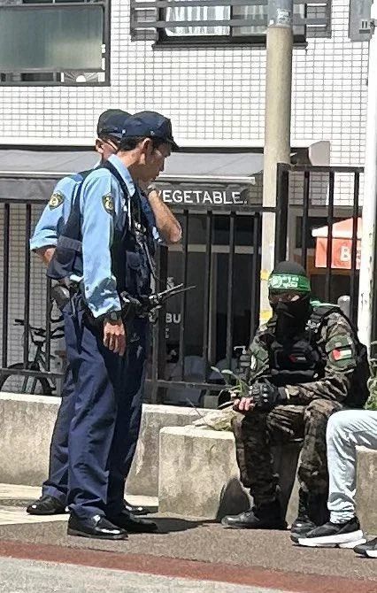 Two police officers stood in front of a guy in full Hamas getup who's sat down. They are presumably placing him under arrest or something idk