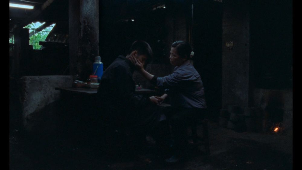 Screenshot: Viet and his mother sitting on chairs next to a table in their very dark house, he's holding her fingers after painting her nails and she cups his cheek in her hand 