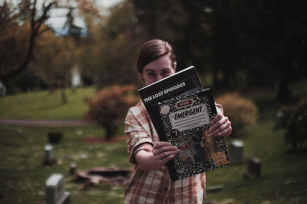 The blurry background of a graveyard. A figure stands in the foreground, wearing a yellow and white flannel with the sleeves rolled up. They have blond hair, and the image is cut off at their waist. They are holding two books straight out in front of them, obscuring most of their face and body, but their eyes can be seen looking over the tops of the book. The second book has only the title visible, which reads "The Lost Episodes." The other book reads "Emergent." It looks like a black and white notebook cover.