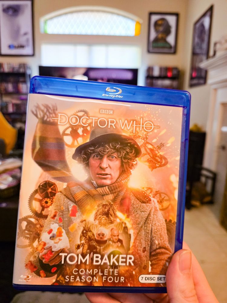 The blu-ray release of the fourth Tom Baker / Fourth Doctor season. It features a orange toned image of the Fourth Doctor clad in a grey coat, scarf flying over his shoulder, jelly babies floating out a pouch. Monsters in front of him. Gears behind him. I'm holding it up in my living room with some movie posters on the walls. 