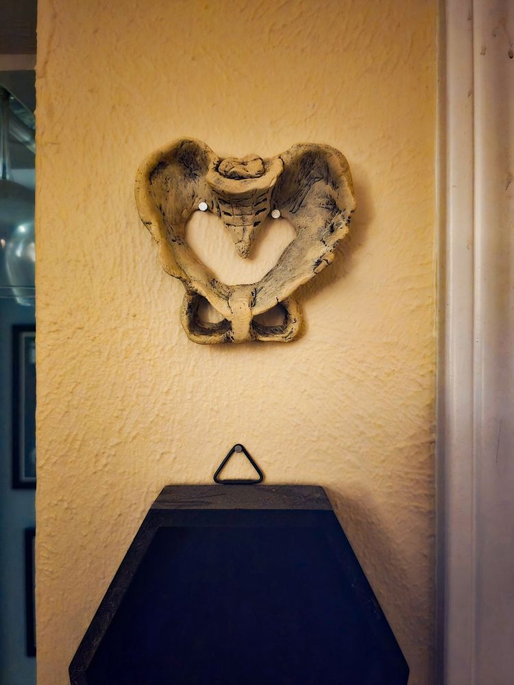 A small sculpture of a human pelvis painted in none with weathering. It is nailed to the wall with two nails. It's mounted over a black coffin. 
