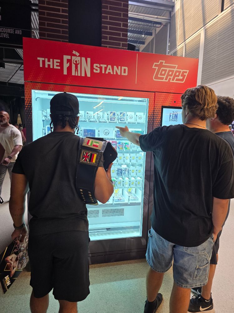 Some guys stand in front of a Topps The Fan Stand baseball card vending machine. One holds a wrestling championship. One points at the cards. The machine is red and has a big clear window. 