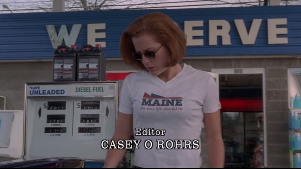 Agent Scully at a gas station. She is wearing a Maine tshirt. Behind her the building says WE SERVE. There is a gas pump to the left of her. 