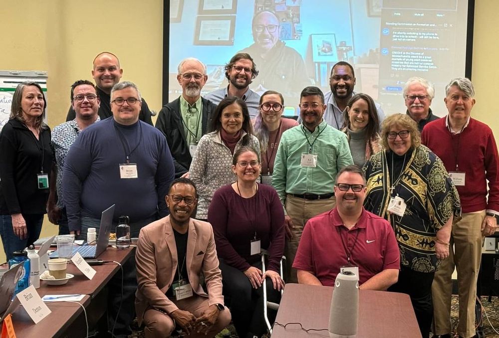 A group photograph of mostly smiling people in a conference room, a mix of all genders and races and adult ages. There is a screen behind the people displaying Zoom participant(s) too.