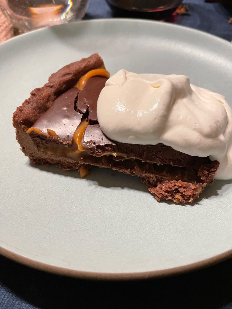Photograph of a slice of caramel chocolate truffle tart with a dollop of cream on a light colored plate 