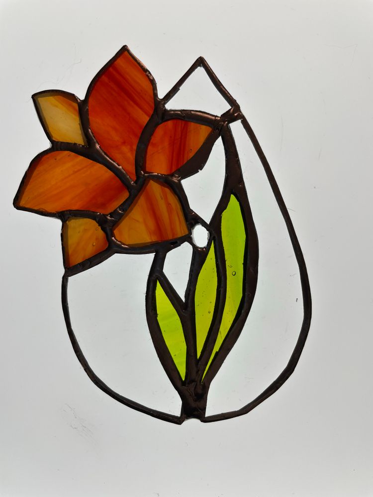Backlit stained glass suncatcher. It’s teardrop shaped and features a red orange lily