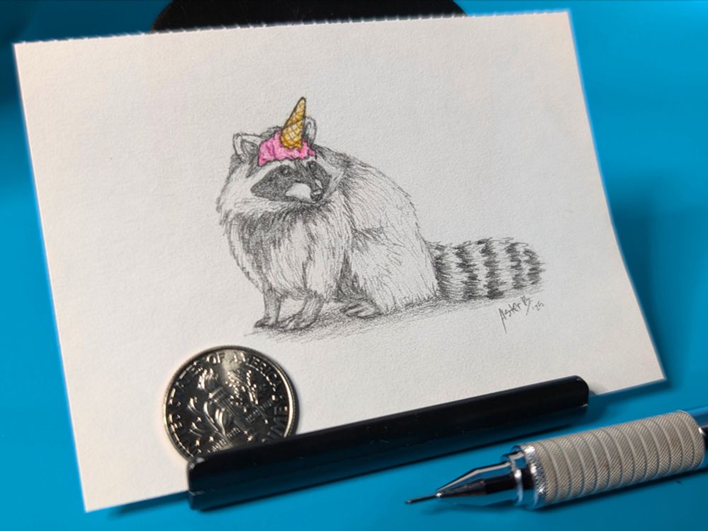 A sketch is propped in a mini easel. It is of a raccoon with an upsidedown ice cream cone on its head, making it resemble a unicorn. 