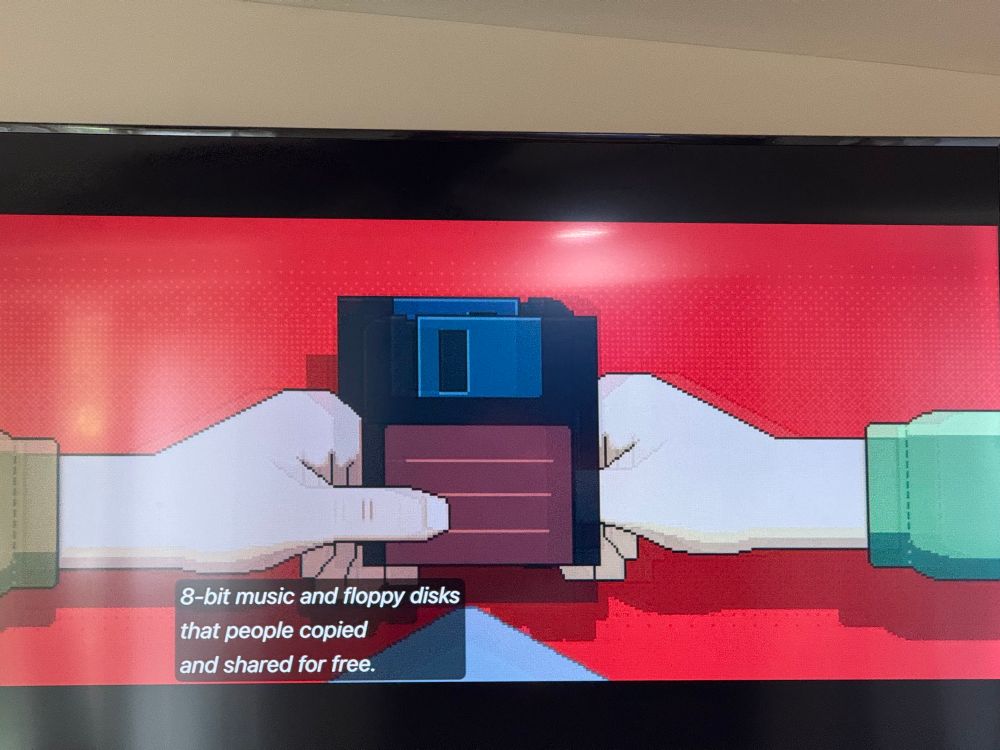 A photo of a television showing a scene from Tetris (2024). Animated hands hold 3.5” floppy disks. The closed captioning reads “8-bit music and floppy disks that people copied and shared for free.”