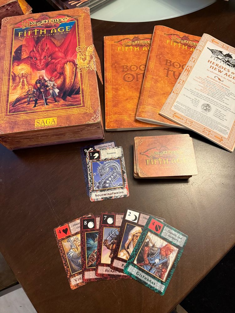 Dragonlance: Fifth Age RPG displayed on a wooden table. Top left is the box which has an image of a red dragon and some adventurers. Top right has the three books that come with the game. A deck of Fifth Age cards is below them.