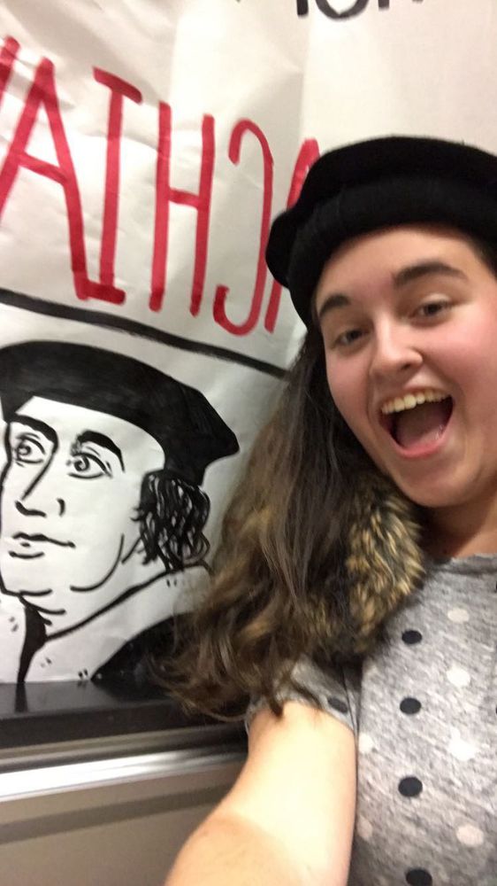 A selfie of a woman in a flat black cap wearing fake fur around her shoulders, taken next to a poster someone drew of a man dressed in the same clothes.