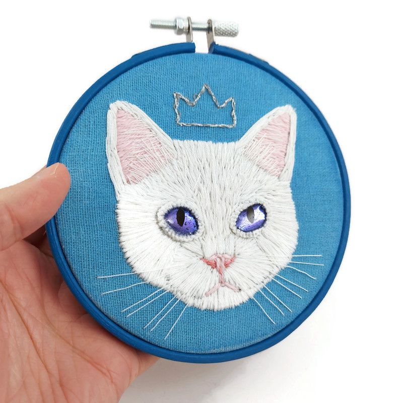 Broderie d'une tête de chat blanc coiffé d'une couronne argentée, réalisée sur une toile bleue ciel tendue sur un cercle à broder en bois bleu.
Les yeux du chat sont deux morceaux de CD fixés à la toile avec la technique de la broderie miroir, puis recouverts de résine époxy teintée bleue foncée.