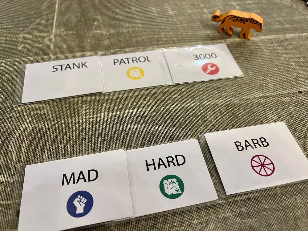 A picture of a card game in play. Each card has a word, a colour, and a symbol on. The first name reads ‘Stank Patrol 3000’ and the second reads ‘Mad Hard Barb’